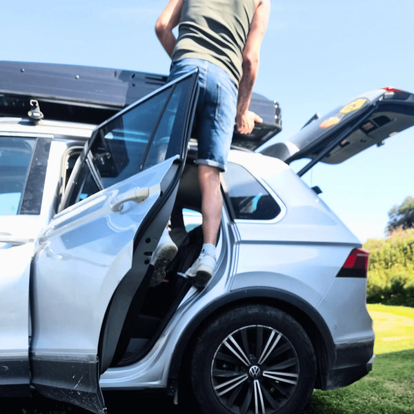 Une personne utilisant une échelle pour accéder à une tente de toit sur un SUV argenté garé par une journée ensoleillée.