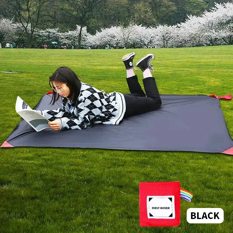 Une personne lisant sur un tapis de poche noir à l'extérieur dans un parc herbeux, mettant en valeur le confort et la portabilité pour le camping et les activités en plein air.