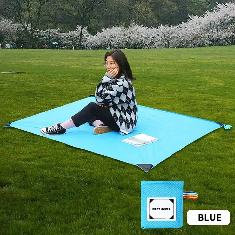 Femme assise sur un tapis de camping bleu en plein air dans un parc, entourée de gazon vert et d'arbres en fleurs, avec une pochette compacte dans le coin.
