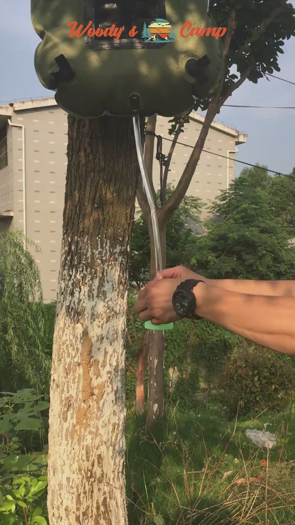 Person using a solar camping shower bag hanging from a tree outdoors.