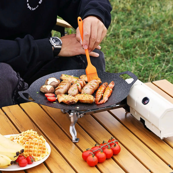 Réchaud de camping portable en utilisation, cuisinant divers aliments en plein air sur une table de pique-nique, mettant en valeur un design polyvalent et compact pour la cuisine en extérieur.