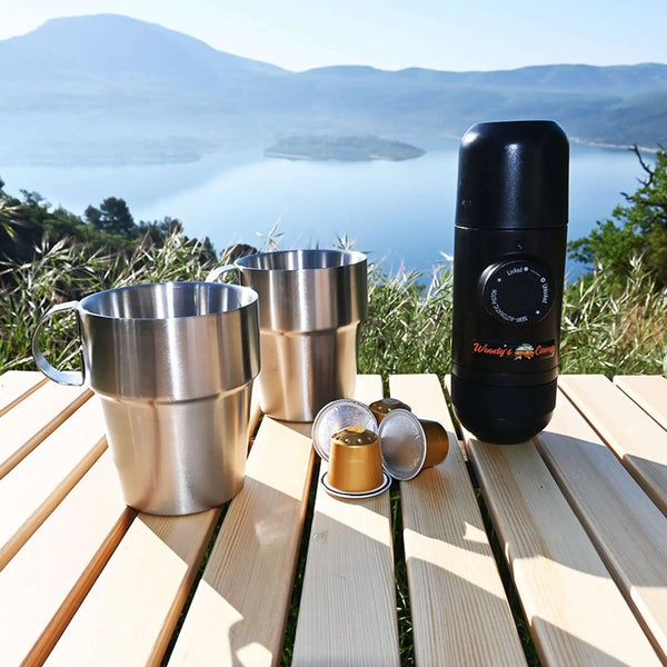 Machine à café Nespresso portable avec des capsules et des mugs sur une table de camping surplombant un lac de montagne pittoresque.
